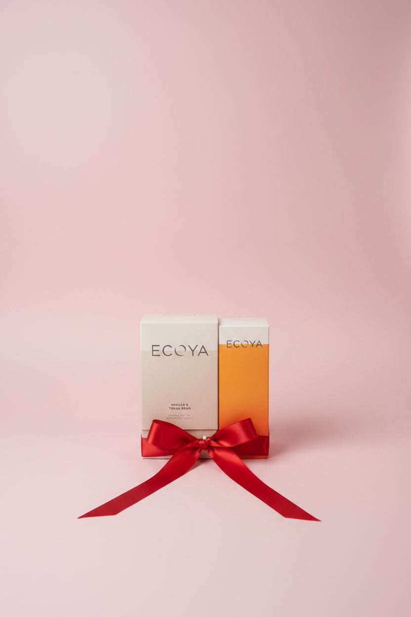 Two ECOYA candles, one vanilla and one orange, with a red bow on a reflective surface.